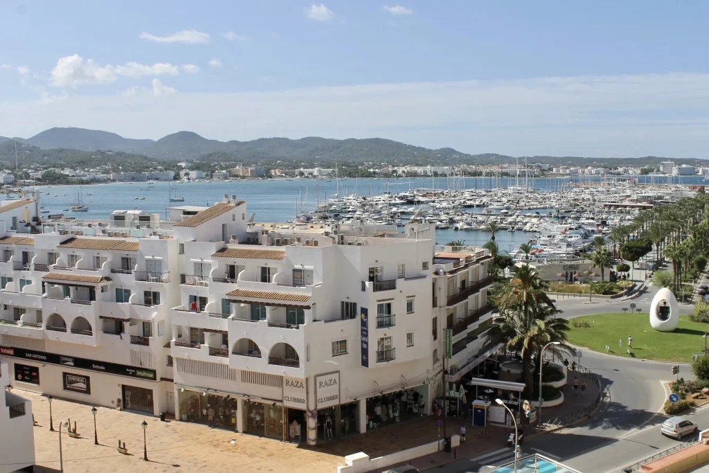 Vista aérea de un puerto deportivo con numerosos barcos y yates en el agua, rodeado de edificios blancos y montañas al fondo bajo un cielo azul.